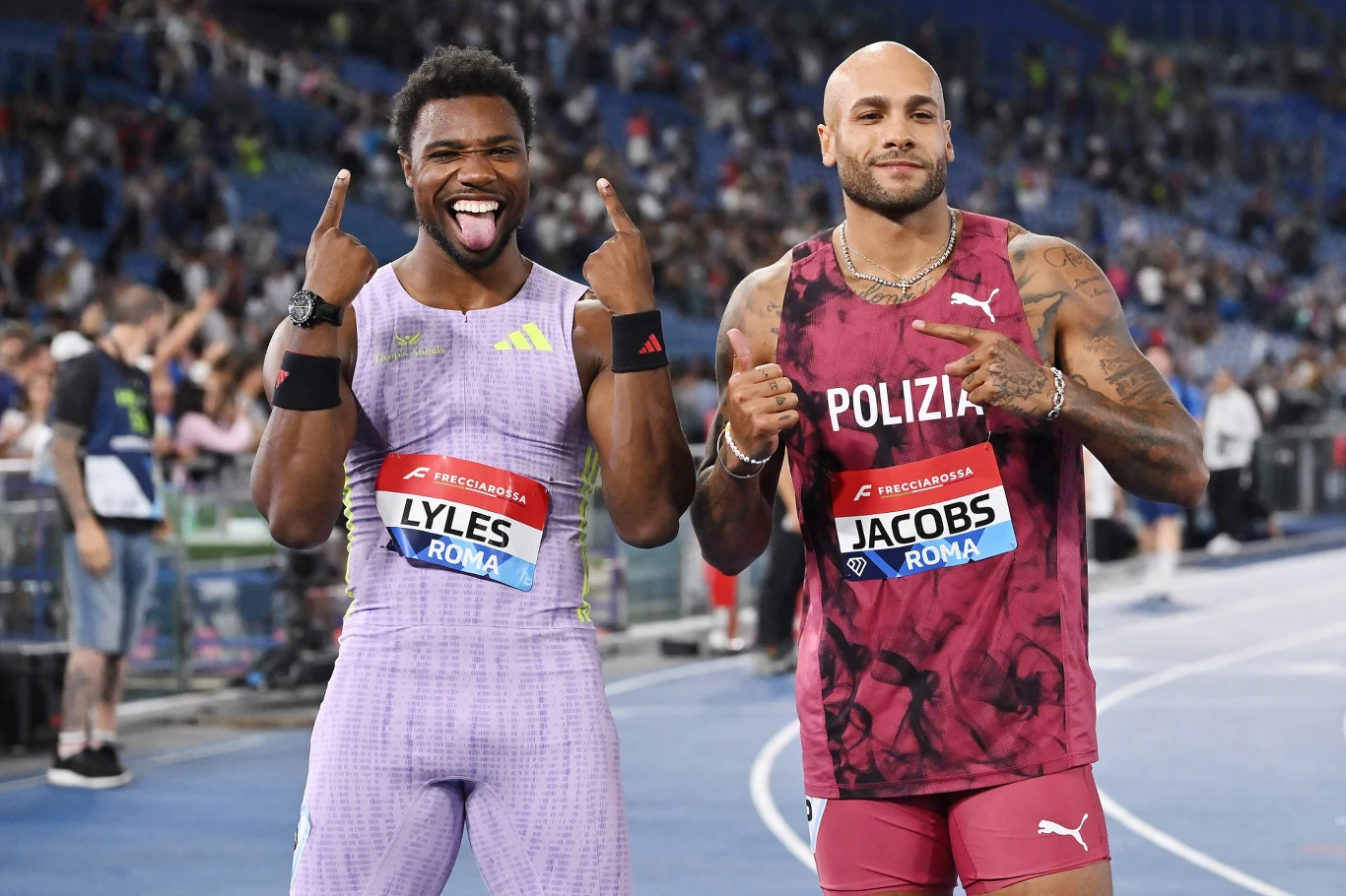 Dwaj lekkoatleci w sportowych strojach stoją obok siebie na bieżni, jeden z nich uśmiechnięty szeroko pokazuje gesty zwycięstwa, drugi prezentuje medal i unosi kciuk do góry.