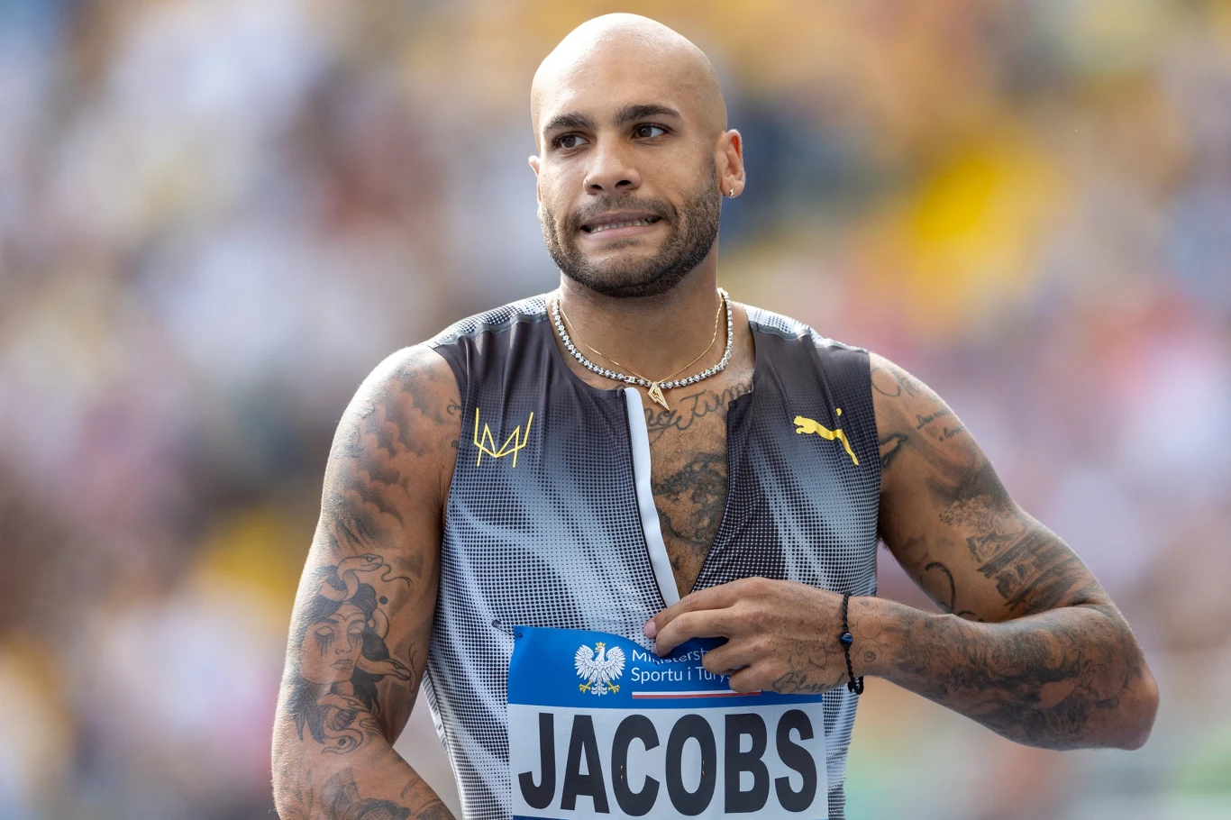 Lekkoatleta z ogolonymi włosami i tatuażami na rękach, ubrany w sportowy strój z nazwiskiem Jacobs oraz numerem startowym, przygotowuje się do biegu na stadionie lekkoatletycznym.