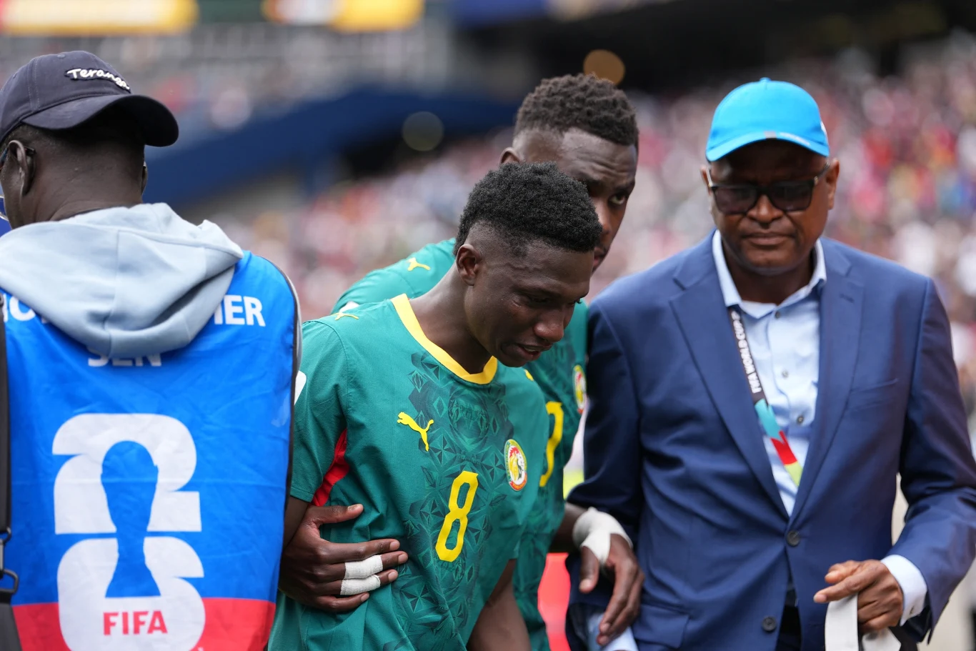 Załamany Lamine Camara Piłkarz w zielonej koszulce z numerem 8 wspierany przez dwóch mężczyzn, jeden w niebieskiej marynarce z identyfikatorem, drugi w odzieży FIFA, podczas wydarzenia sportowego na stadionie.