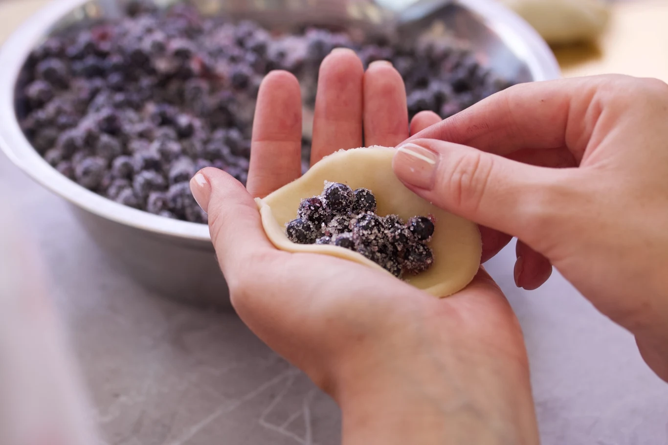 Jeden dodatek do nadzienia i jagody nie wypłyną z pierogów Ręce zawijające pieroga z nadzieniem jagodowym, obok miska z jagodami obtoczonymi w cukrze.