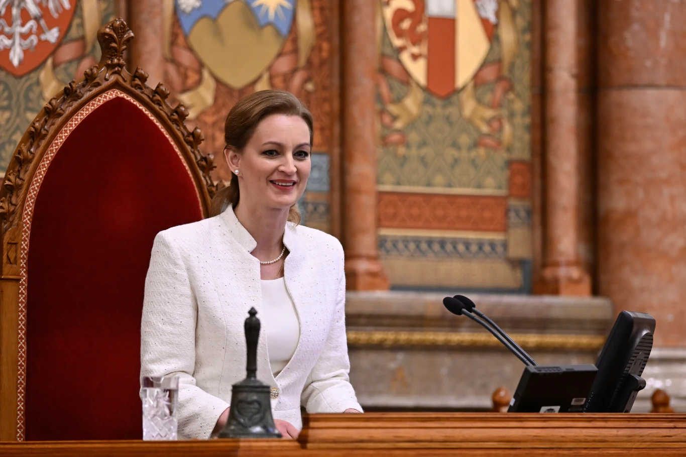 Kobieta w białym eleganckim stroju siedzi na ozdobionym, czerwonym fotelu za drewnianym biurkiem, obok znajduje się mikrofon oraz monitor, w tle widoczne są bogato zdobione elementy architektoniczne i herby.