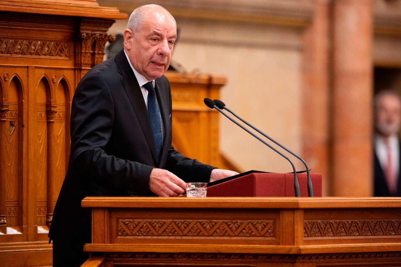Mężczyzna w garniturze przemawia przy drewnianej mównicy z dwoma mikrofonami, po prawej stronie leży szklanka wody.