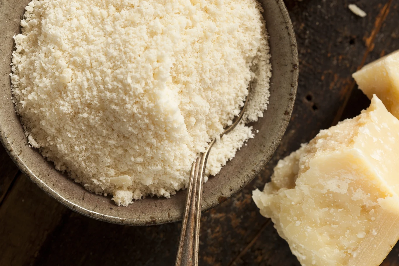 Parmezan, skyr i kefir to produkty mleczne, które mogą wzbogacić codzienną dietę w białko, wapń i składniki wspierające jelita Starte na drobno sery w misce z łyżką oraz kawałki twardego sera na drewnianym blacie.