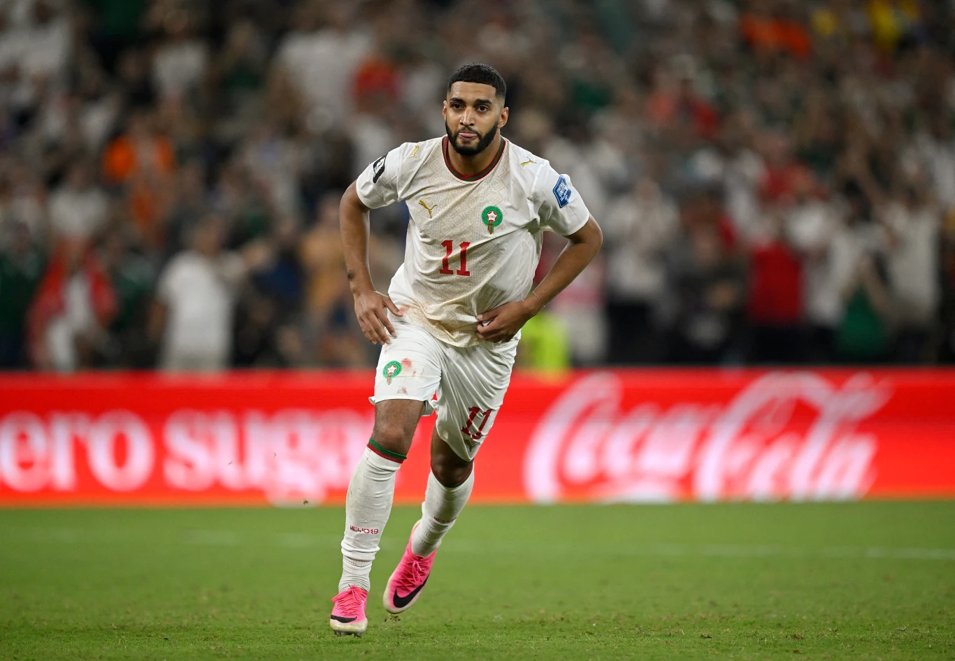 Ismael Saibari Piłkarz w białym stroju reprezentacji Maroka z numerem 11 biegnący po murawie stadionu podczas meczu, z wyraźnie widocznym logo Coca-Cola na tle trybun.