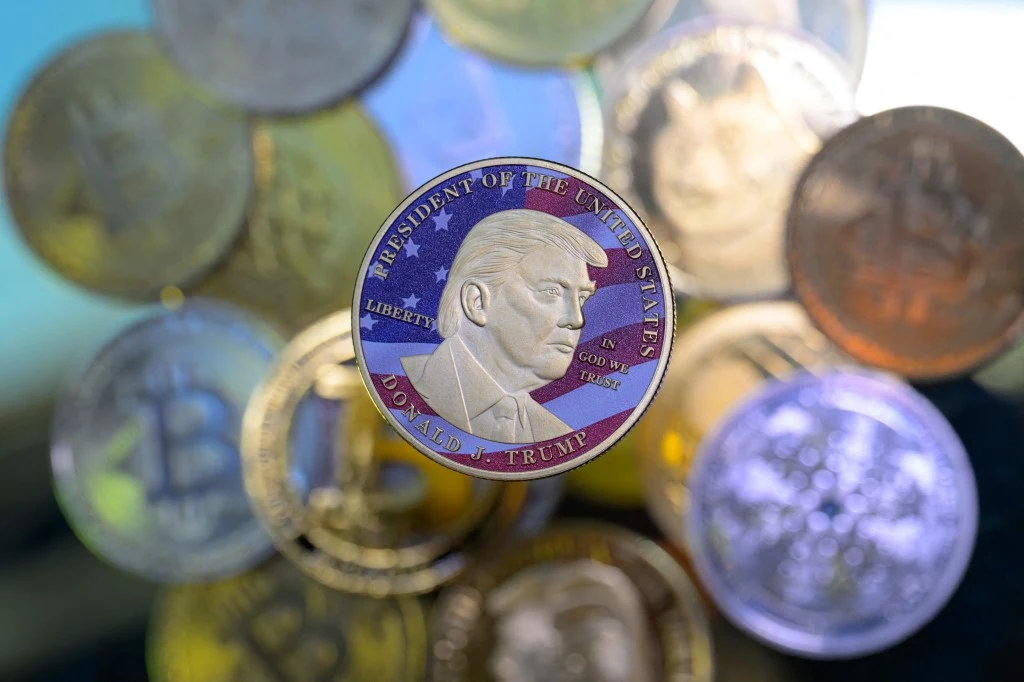 Moneta z wizerunkiem Donalda Trumpa oraz napisem "President of the United States" otoczona innymi kolorowymi monetami o różnych wzorach, w tym z symbolem Bitcoina.