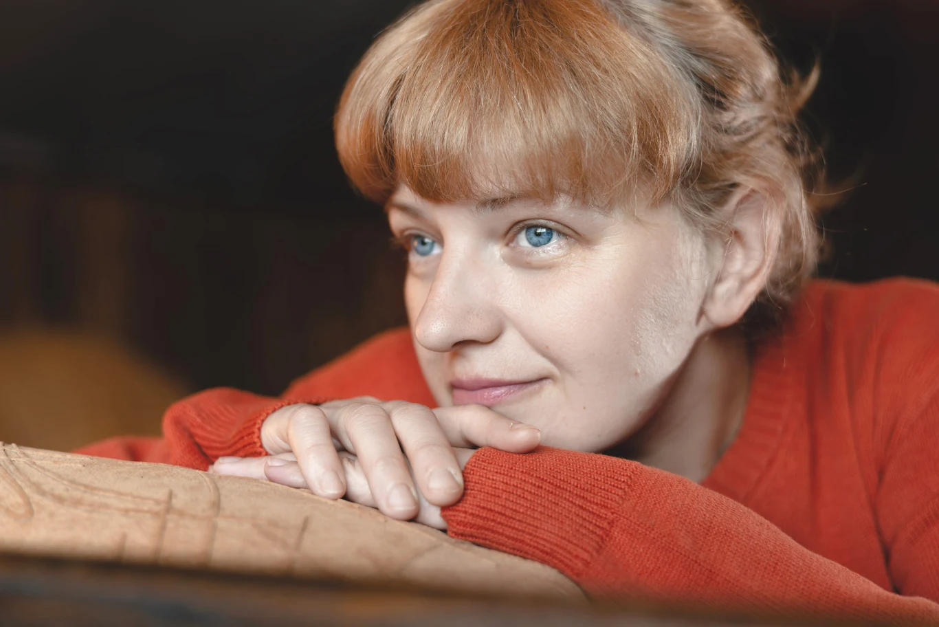 Kobieta o jasnych włosach i niebieskich oczach oparta o kanapę, ubrana w czerwony sweter, patrzy z lekkim uśmiechem w dal.