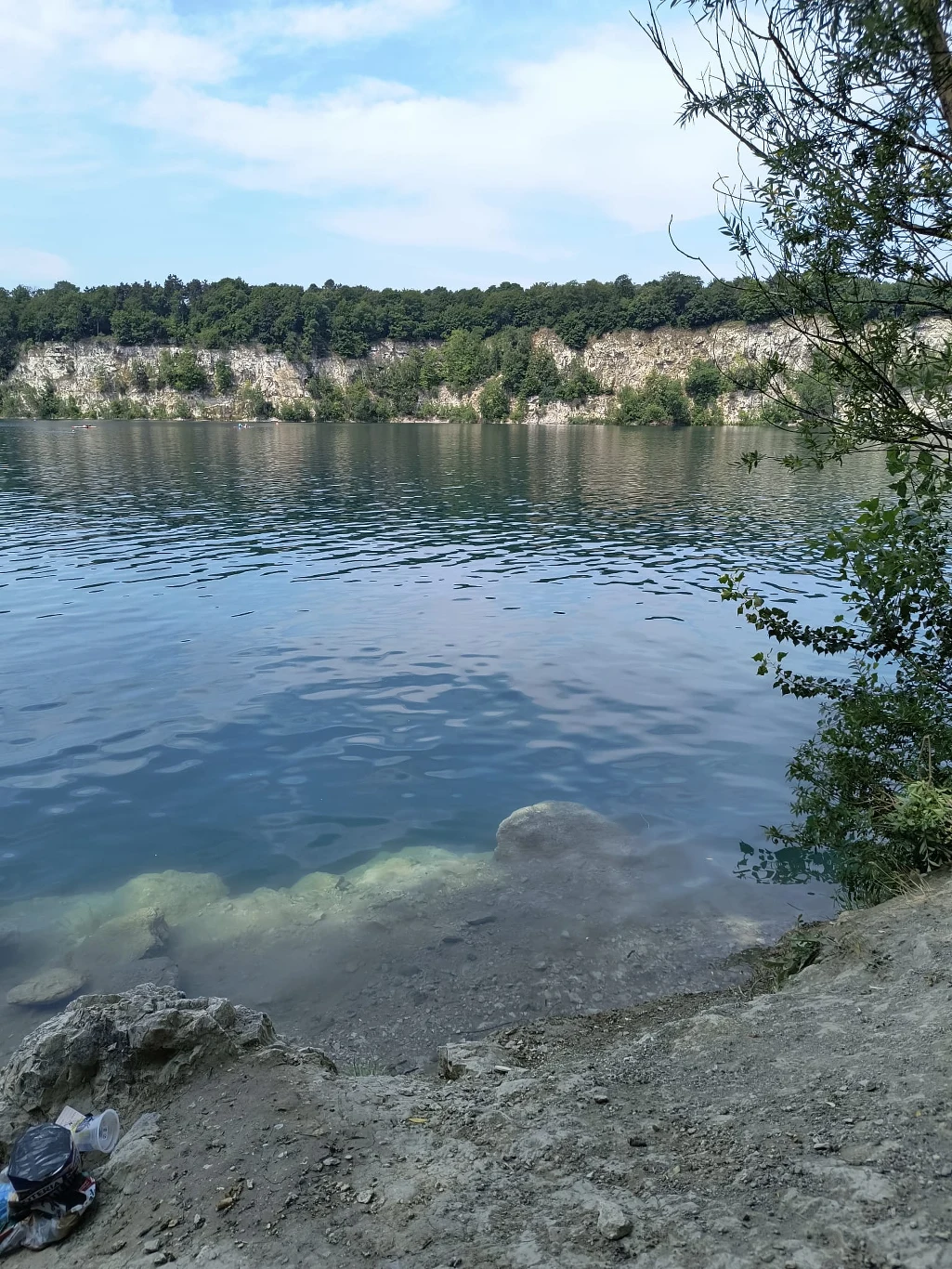 Zakrzówek Spokojna tafla jeziora otoczona skalistym brzegiem i lasem, z niewielkim fragmentem kamienistego wybrzeża na pierwszym planie.