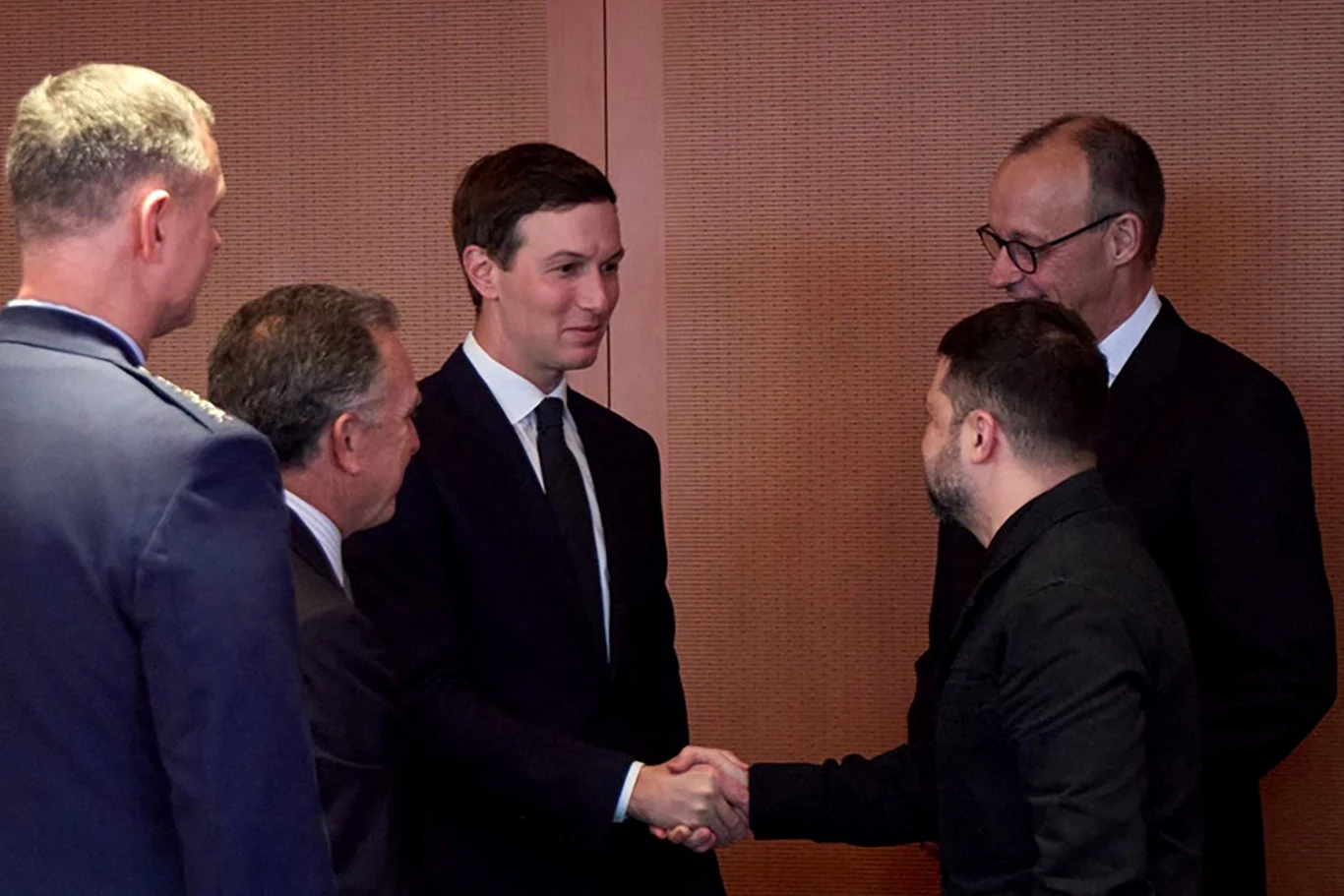 Grupa pięciu mężczyzn w eleganckich garniturach, dwóch z nich ściska sobie dłonie w geście powitania, pozostali przyglądają się wymianie uścisku.