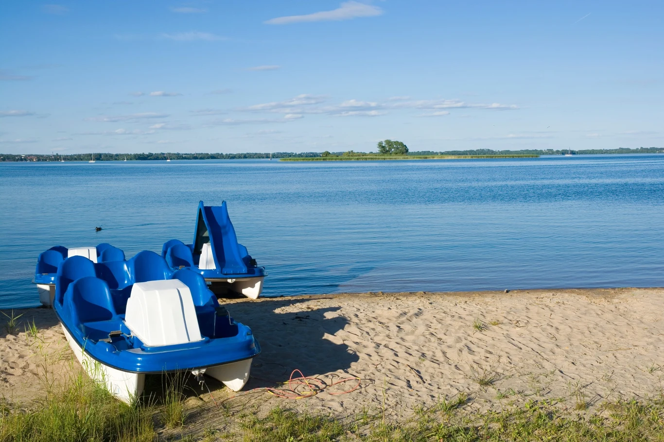 Gdzie warto wybrać się nad wodę blisko Rzeszowa? (zdj. poglądowe) Dwa niebiesko-białe rowery wodne na piaszczystej plaży przy jeziorze, w tle zielona wyspa