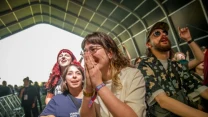 Grupa młodych osób blisko barierek festiwalowej sceny, wyrażających entuzjazm i radość podczas koncertu, jedna z dziewczyn ma splecione ręce przy twarzy, obok niej osoby śmieją się i klaszczą, w tle tłum zgromadzony pod zadaszeniem.