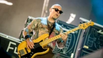 Łysy mężczyzna w okularach przeciwsłonecznych, z licznymi tatuażami, gra na elektrycznej gitarze basowej na scenie podczas koncertu, w tle widoczne głośniki wzmacniaczy.