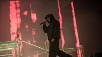 Raper w ciemnej bluzie z kapturem trzyma mikrofon i występuje na scenie z industrialną konstrukcją, w tle pionowe czerwone światła.