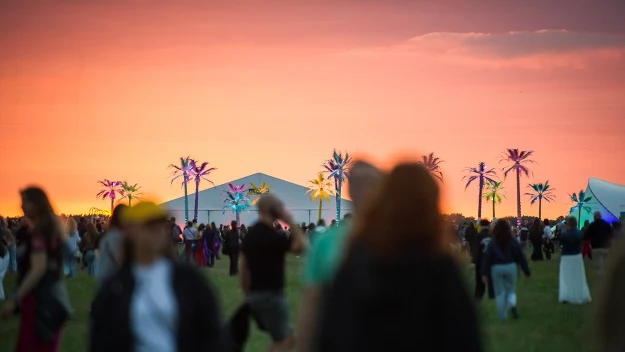 Grupa ludzi na festiwalu na otwartej przestrzeni, kolorowe sztuczne palmy i namiot sceniczny na tle pomarańczowego nieba o zachodzie słońca.