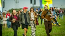 Grupa młodych osób spaceruje po trawie podczas plenerowej imprezy, ubrani w swobodne, kolorowe stylizacje, rozmawiają i trzymają napoje w dłoniach.