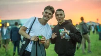 Dwóch młodych mężczyzn stoi razem na trawie z napojami w dłoniach, uśmiechając się i pozując do zdjęcia podczas plenerowej imprezy o zachodzie słońca.