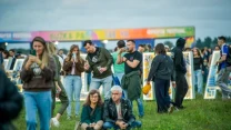 Grupa ludzi bawiących się na festiwalu plenerowym, niektórzy siedzą na trawie, inni stoją lub rozmawiają, w tle kolorowe stoiska i festiwalowe dekoracje.
