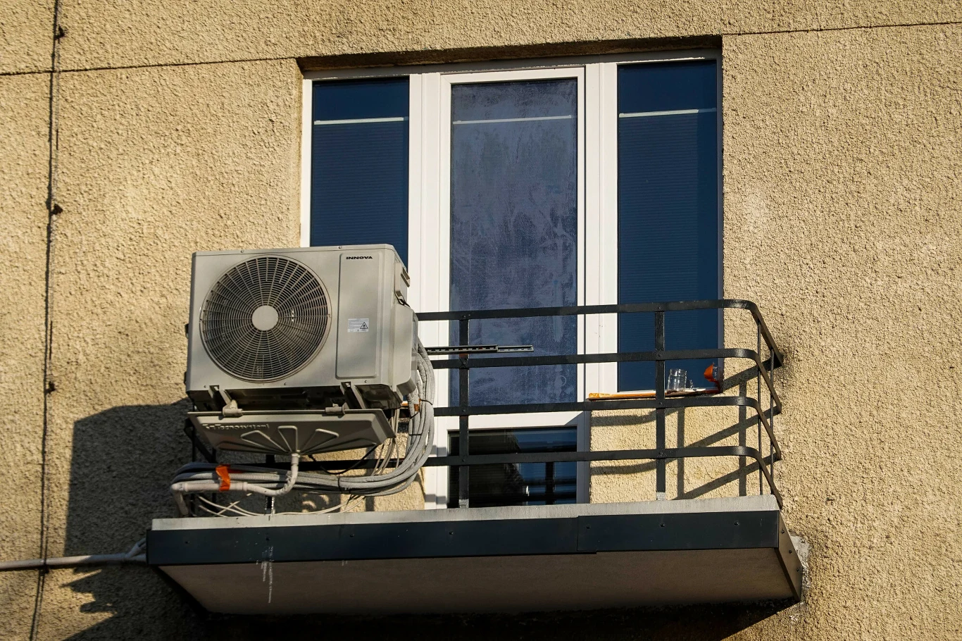 Klimatyzator zamontowany na małym balkonie przed białymi drzwiami balkonowymi w budynku o żółtej elewacji.
