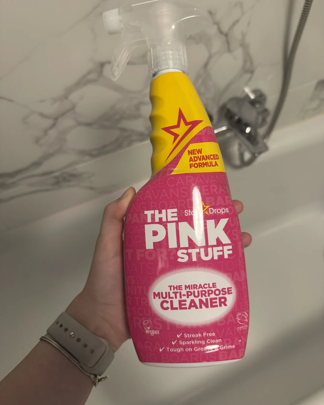 Ręka trzyma butelkę środka czyszczącego The Pink Stuff z żółto-różową etykietą na tle białej wanny i baterii łazienkowej.
