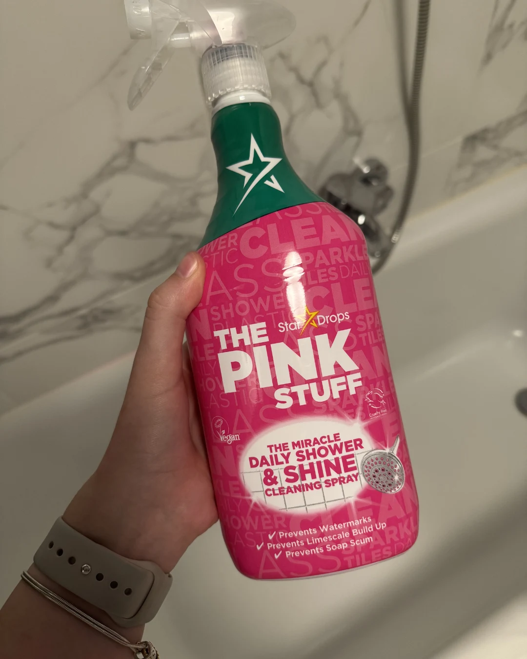 Różowa butelka środka czyszczącego The Pink Stuff z białym spryskiwaczem trzymana w lewej ręce na tle białej wanny oraz baterii łazienkowej ze słuchawką prysznicową.
