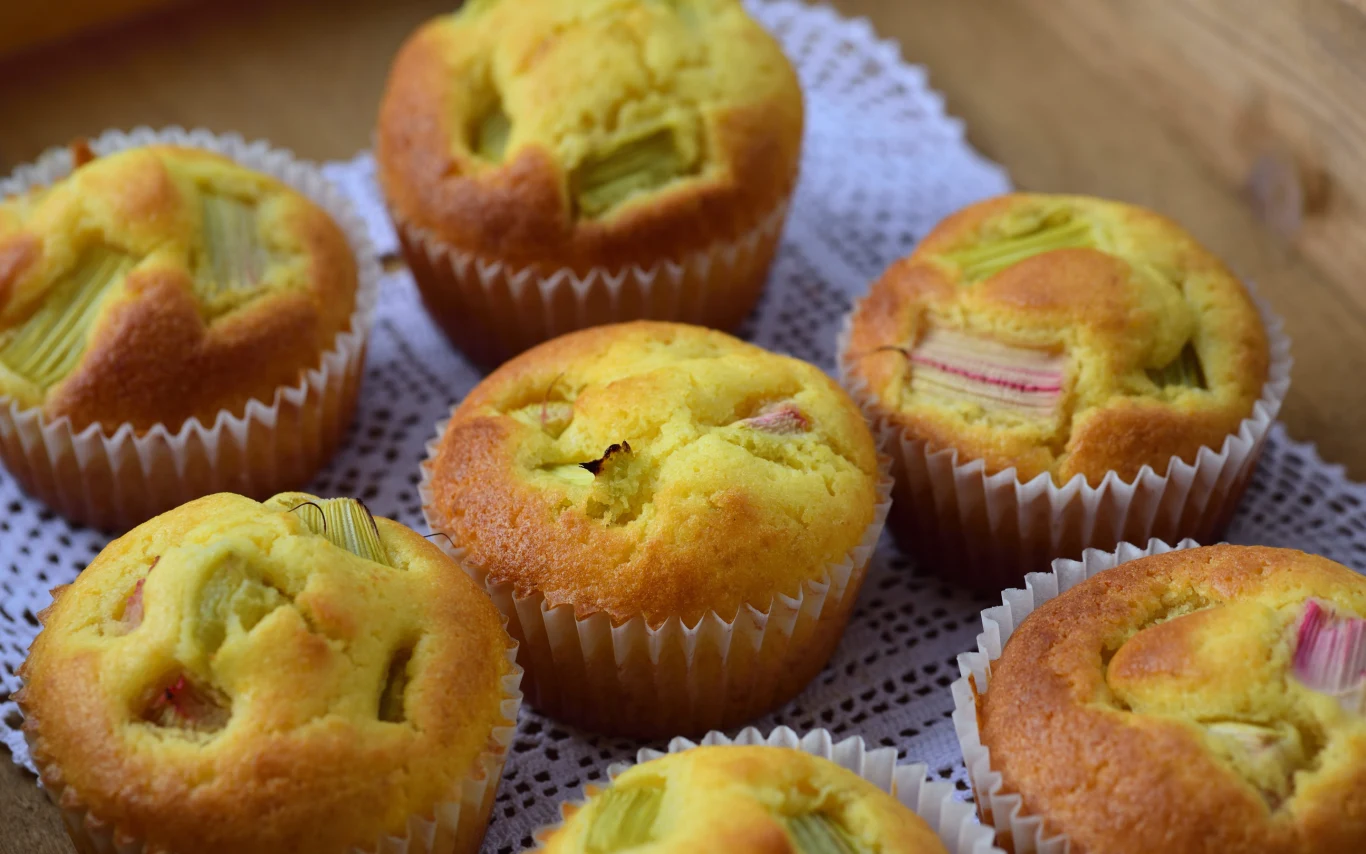 Kwaśny rabarbar świetnie komponuje się ze słodkim ciastem na muffinki Siedem muffinek z rabarbarem na białej serwetce, widoczne kawałki rabarbaru w cieście.