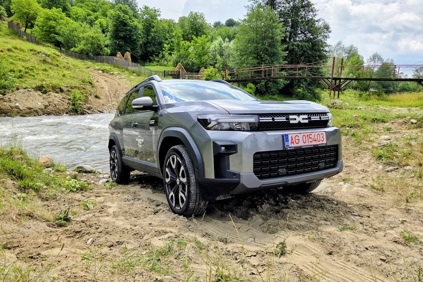 Nowoczesny SUV marki Dacia zaparkowany na piaszczystym brzegu rzeki, otoczony zielenią i naturalnym krajobrazem, w tle widoczny most drewniany oraz bujna roślinność.