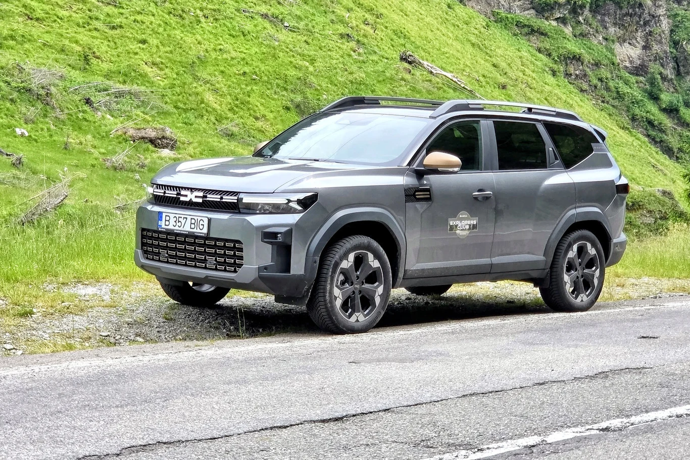 Szary SUV marki Dacia zaparkowany przy drodze asfaltowej, w otoczeniu zielonych, trawiastych wzgórz.