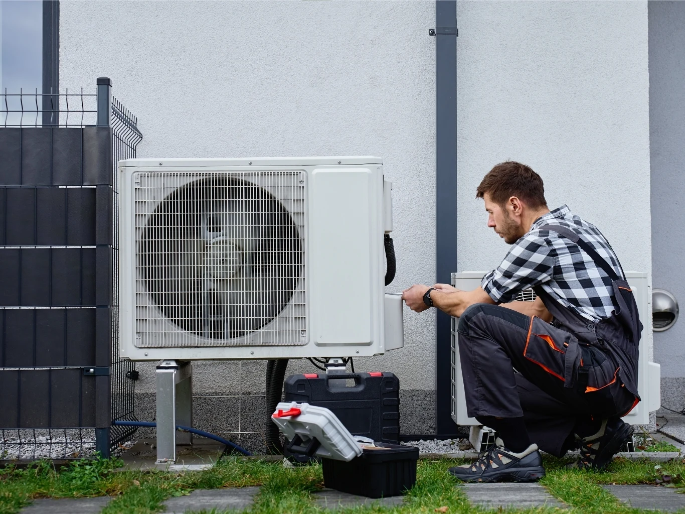 Montaż klimatyzacji na elewacji bloku mieszkalnego wymaga specjalnego pozwolenia od wspólnoty Mężczyzna w roboczym ubraniu wykonuje naprawę lub przegląd jednostki zewnętrznej pompy ciepła przy budynku, obok niego otwarta skrzynka z narzędziami.