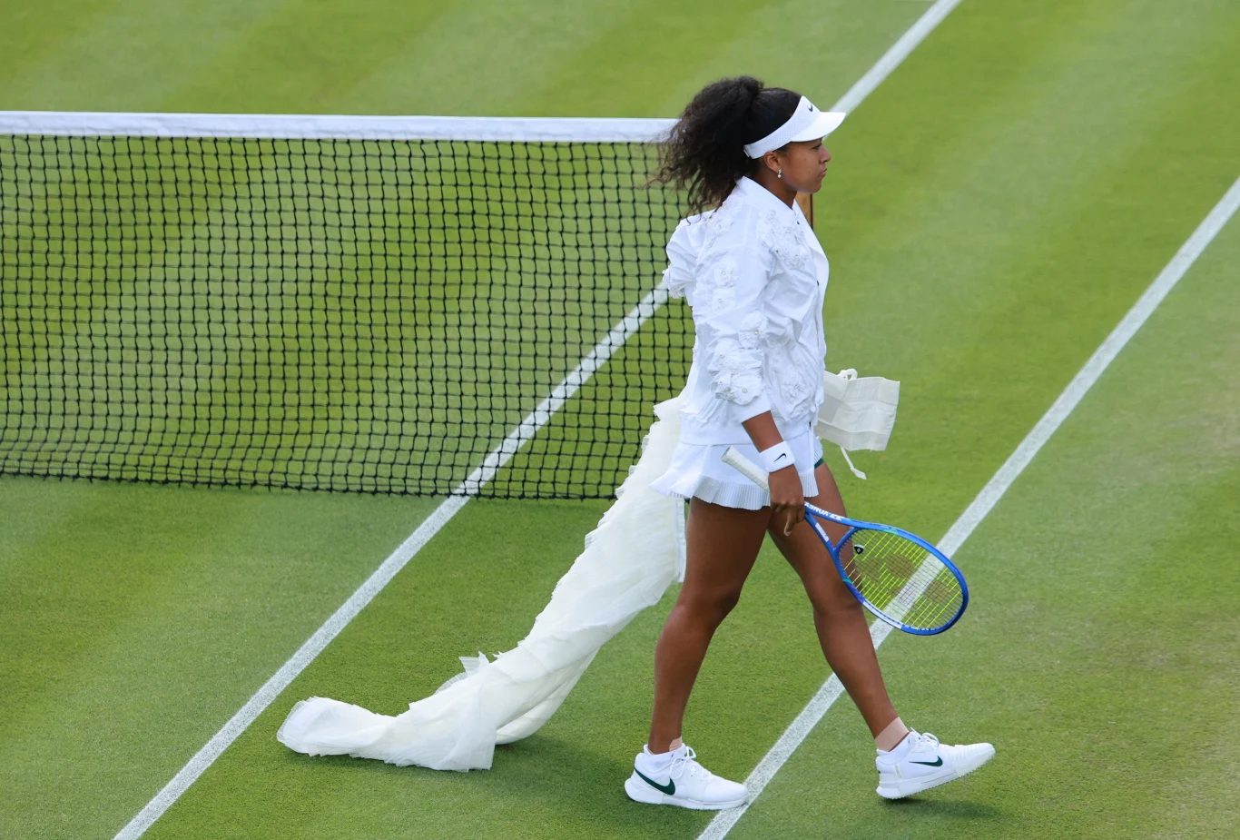 Naomi Osaka Tenisistka w białym stroju z długim, efektownym trenem przechodzi obok siatki na kortcie trawiastym, trzymając rakietę tenisową w prawej ręce.