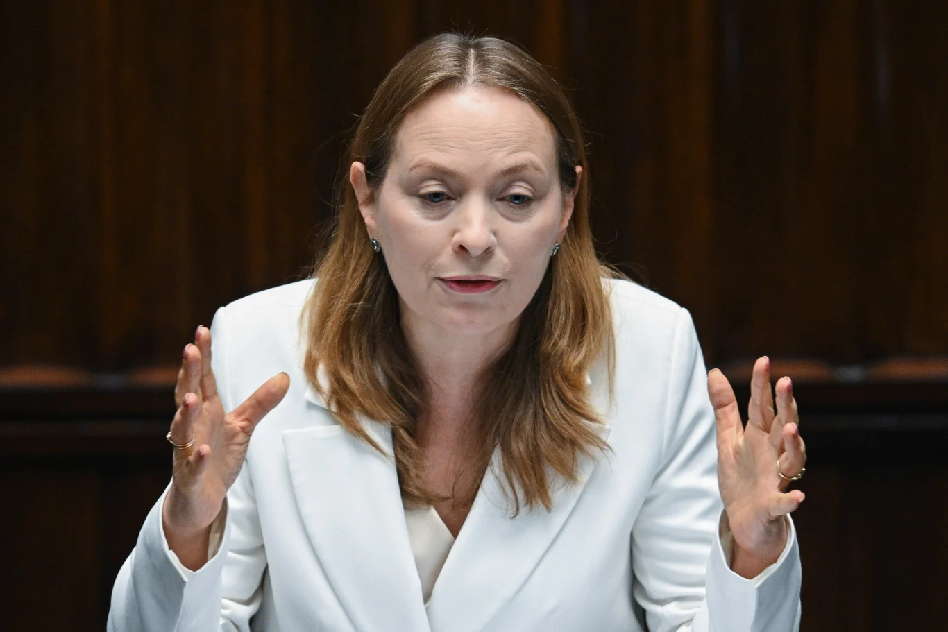 Minister funduszy i polityki regionalnej Katarzyna Pełczyńska-Nałęcz Katarzyna Pełczyńska-Nałęcz w białym żakiecie gestykuluje na tle ciemnego tła podczas wypowiedzi.