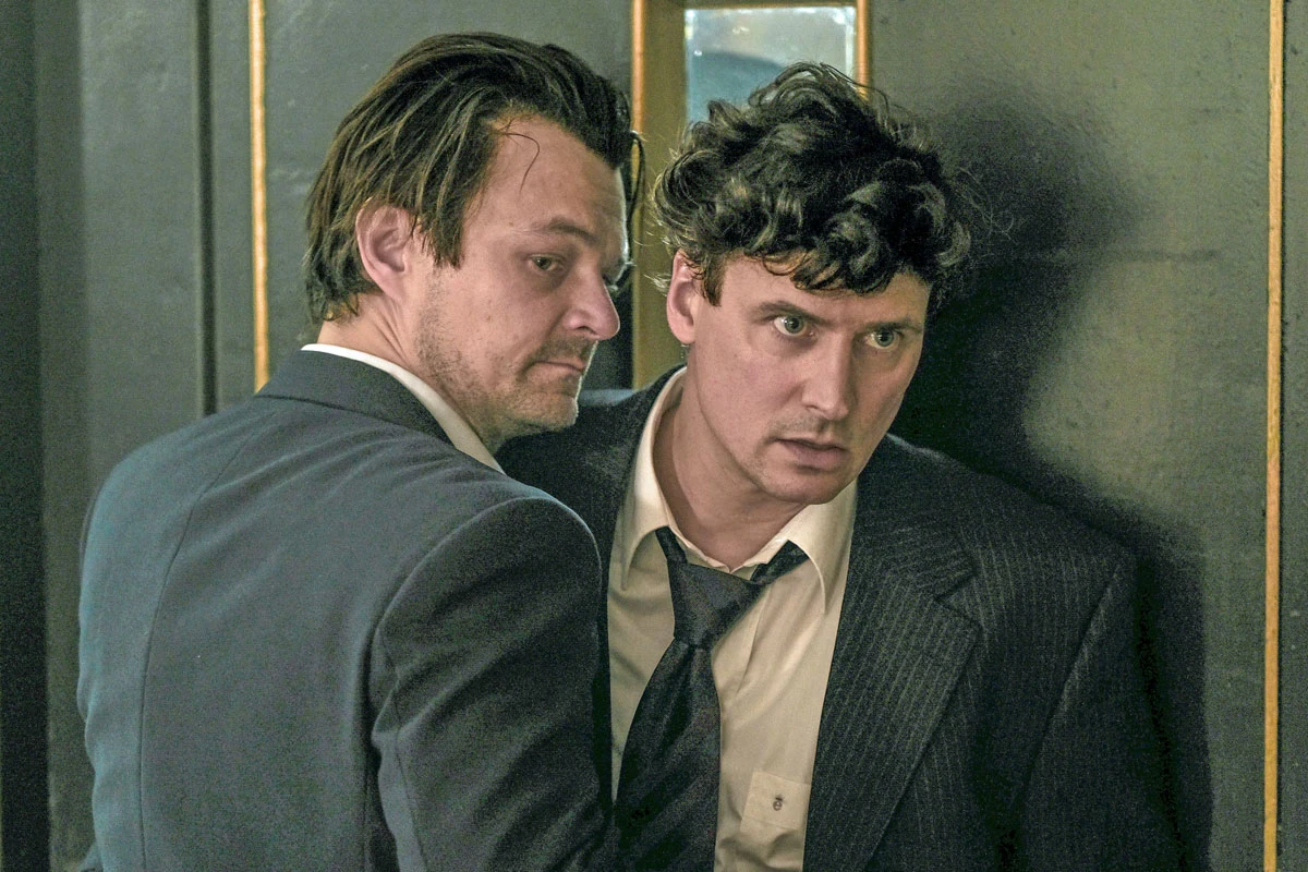 Dawid Ogrodnik i Mateusz Damięcki na planie filmu "Zdrada" Dawid Ogrodnik i Mateusz Damięcki w garniturach na planie filmu "Zdrada", stojący przy drzwiach.