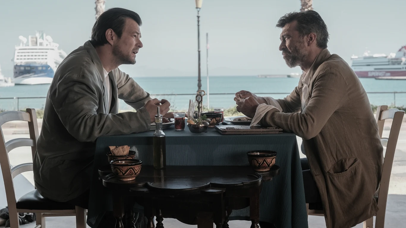 Dawid Ogrodnik i Marcin Czarnik na planie "Zdrady" Dwóch mężczyzn siedzi naprzeciwko siebie przy stole w restauracji z widokiem na port i zacumowane statki, rozmawiając podczas posiłku.