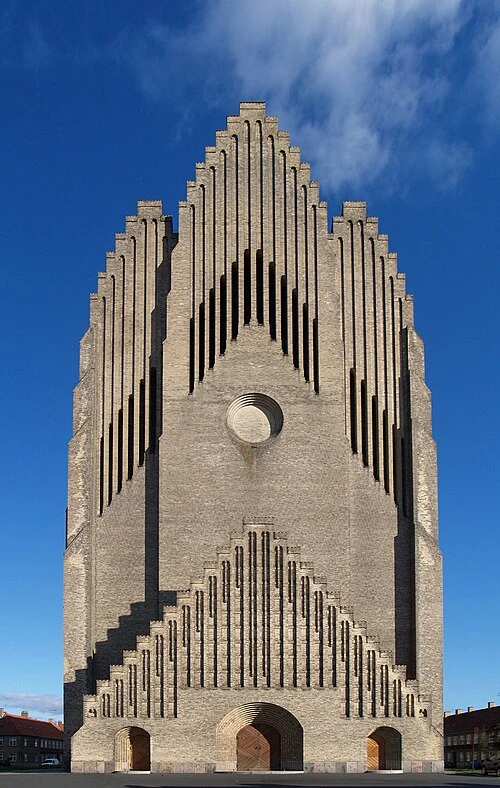 Monumentalna fasada kościoła o unikalnej geometrycznej formie z jasnych cegieł, z wyraźnymi pionowymi podziałami i charakterystycznymi schodkowymi zdobieniami wokół dużego okrągłego okna.