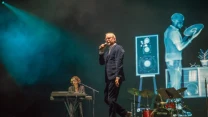 Wokalista w garniturze śpiewa na scenie, z tyłu klawiszowiec przy instrumencie oraz perkusista przy zestawie perkusyjnym, na ekranie za nimi wyświetla się wizualizacja z postacią, głośnikiem i rośliną.