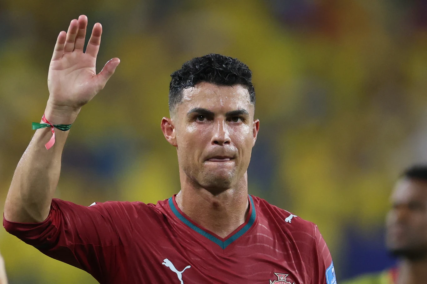 Cristiano Ronaldo Piłkarz w bordowej koszulce z herbem Portugalii unosi prawą rękę w górę, patrząc przed siebie, na tle rozmytych postaci i żółtego stadionowego tłumu.