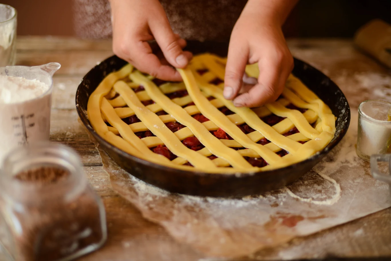 Crostata jest prosta w przygotowaniu Ręce układające paski ciasta kruchego na wierzchu okrągłej formy wypełnionej owocowym nadzieniem, na drewnianym blacie pokrytym mąką.