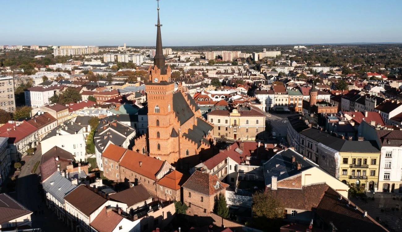 Ceglany kościół z wysoką wieżą otoczony zwartą zabudową niskich kamienic i domów na tle rozległego miasta.