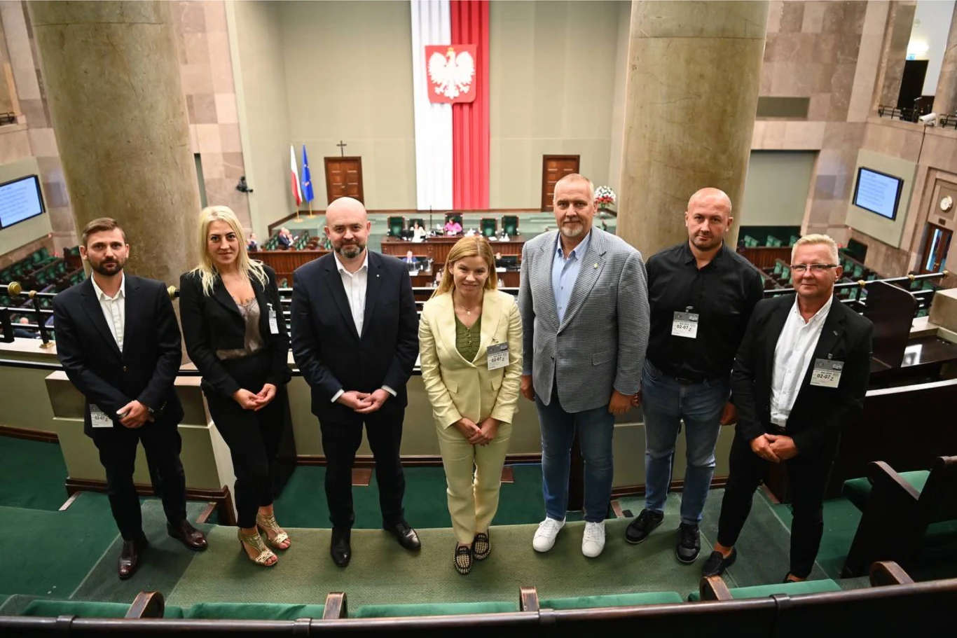 Grupa siedmiu osób stojących w pierwszym rzędzie na tle sali plenarnej polskiego parlamentu z widoczną flagą narodową i godłem Polski na ścianie głównej.