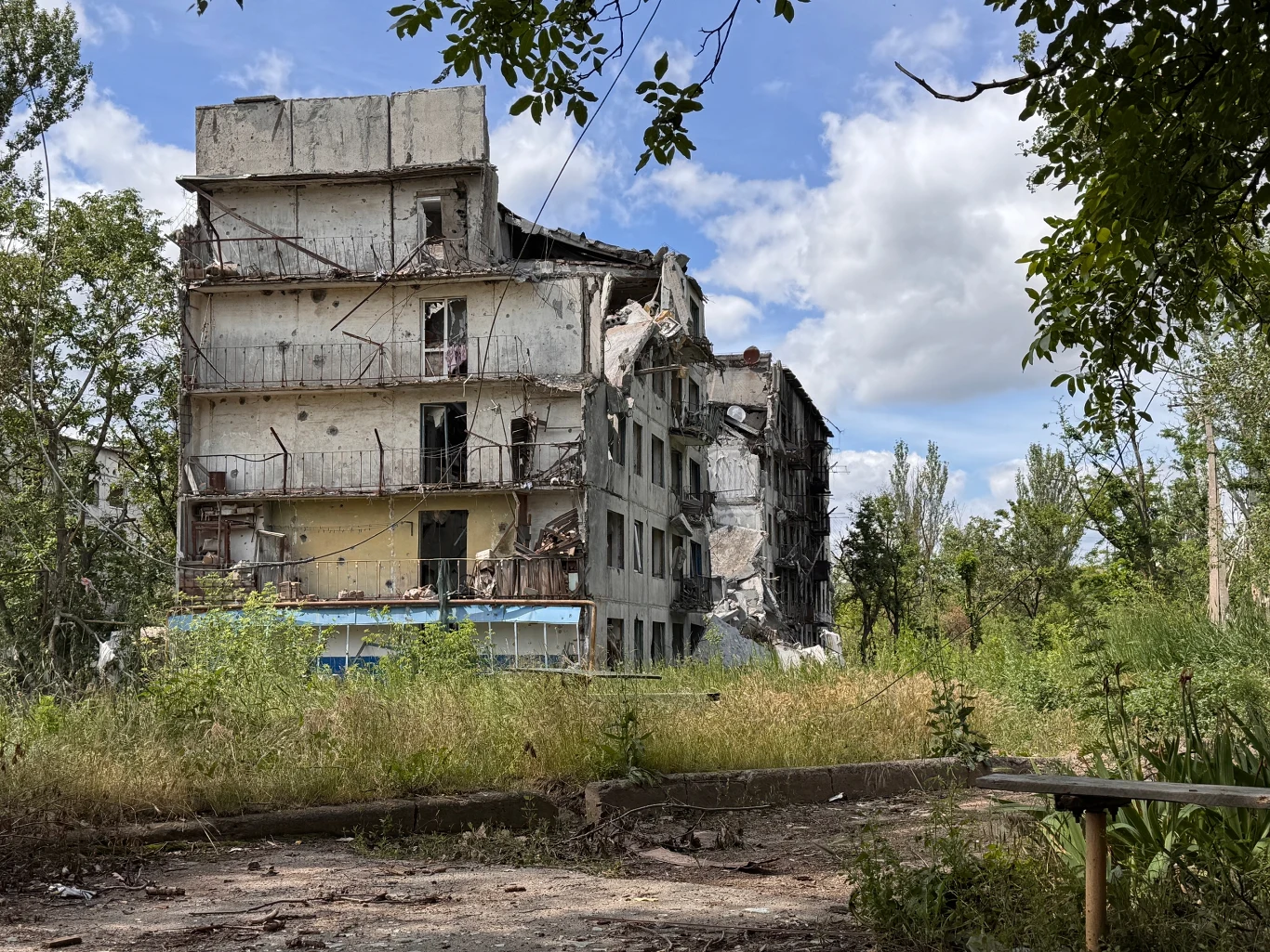 Zniczony budynek w miejscowości Oleksijewo-Drużkiwka, niedaleko od KOnstantynówki Zniszczony wielopiętrowy budynek z licznymi śladami uszkodzeń, otoczony przez roślinność i zaniedbane podwórze.