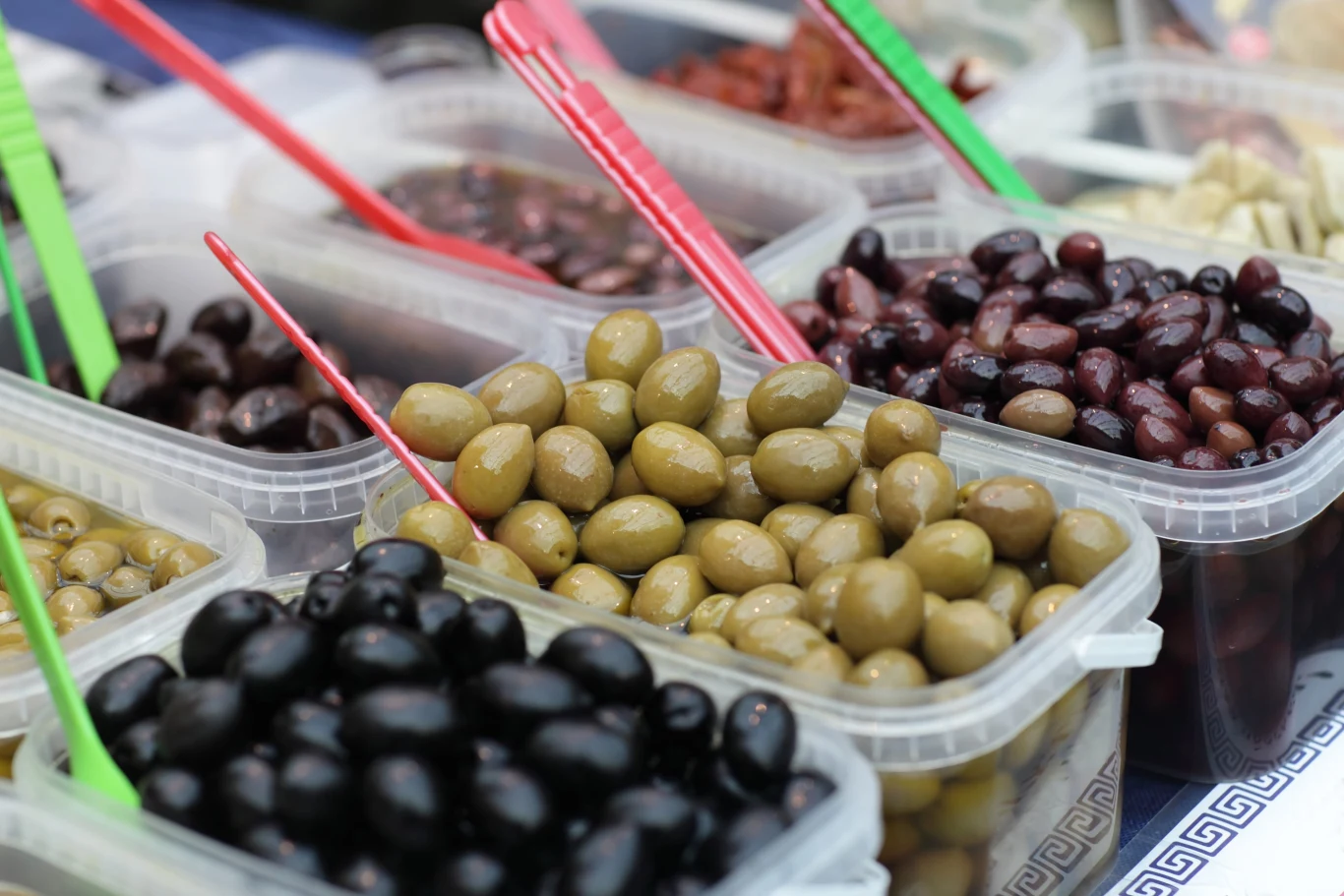 Różne rodzaje oliwek w plastikowych pojemnikach, każdy z kolorową łopatką do nakładania.