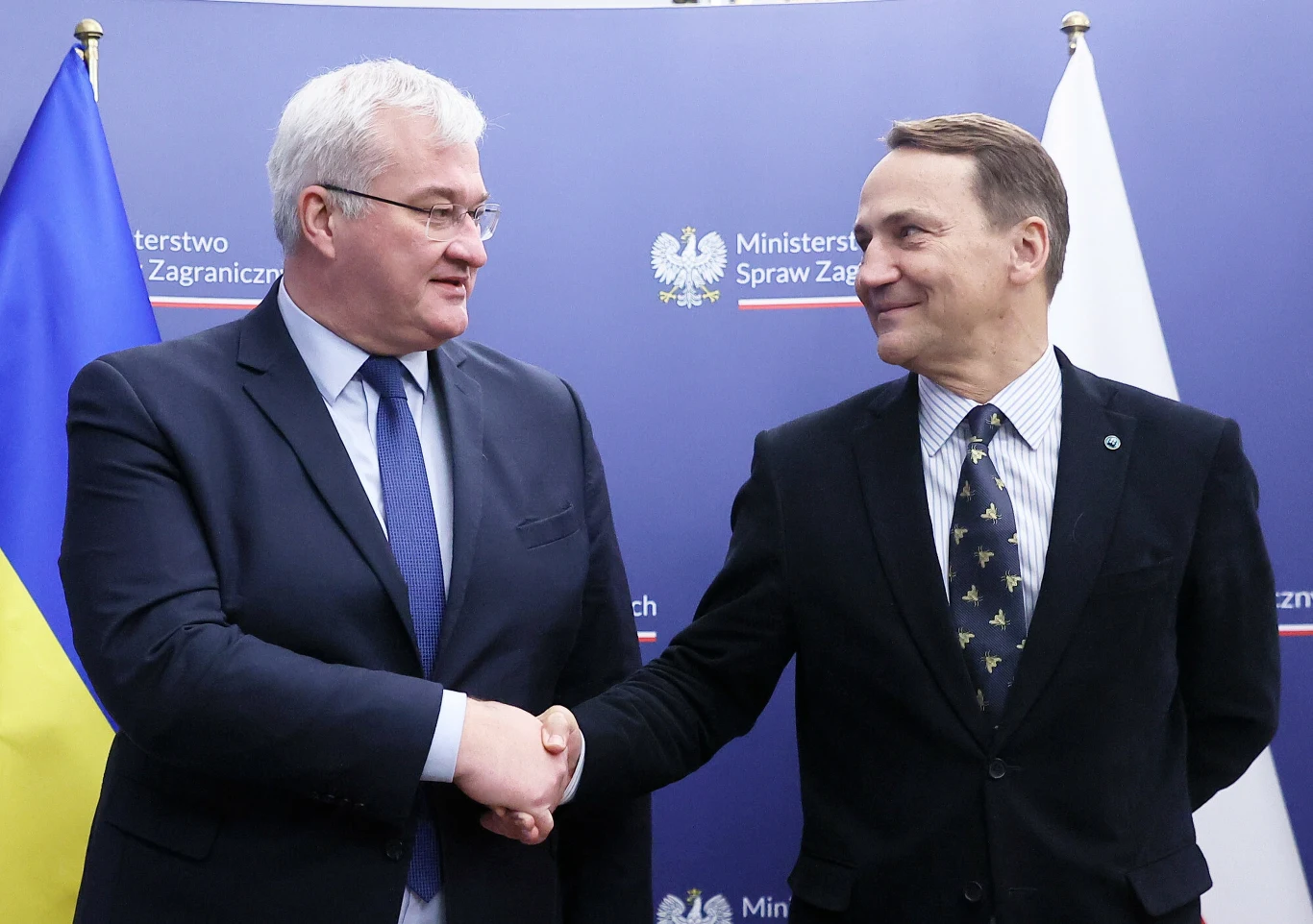 Radosław Sikorski i Andrij Sybiha Dwóch elegancko ubranych mężczyzn podaje sobie ręce na tle flag Ukrainy i Polski oraz tablicy z nazwą Ministerstwa Spraw Zagranicznych.