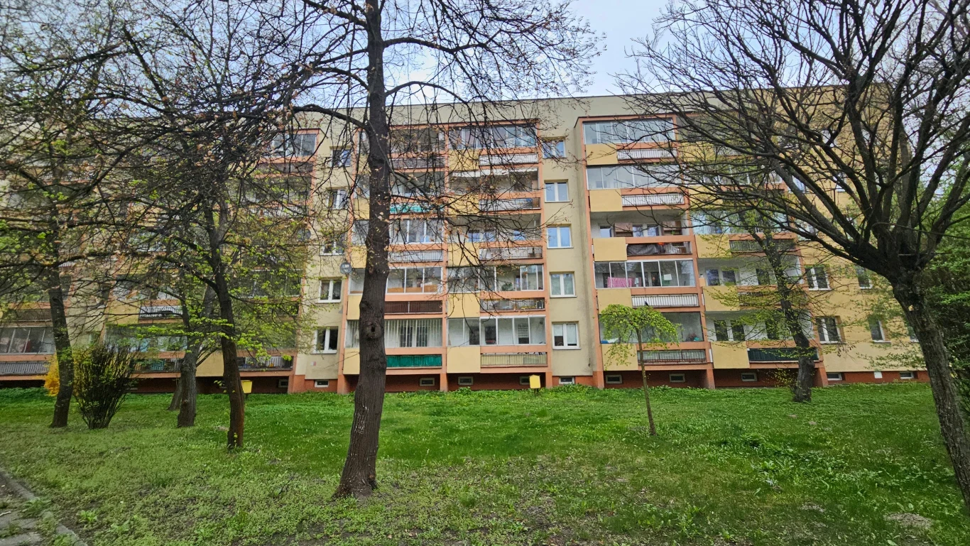 Czteropiętrowy blok mieszkalny z balkonami, otoczony drzewami i trawnikiem.