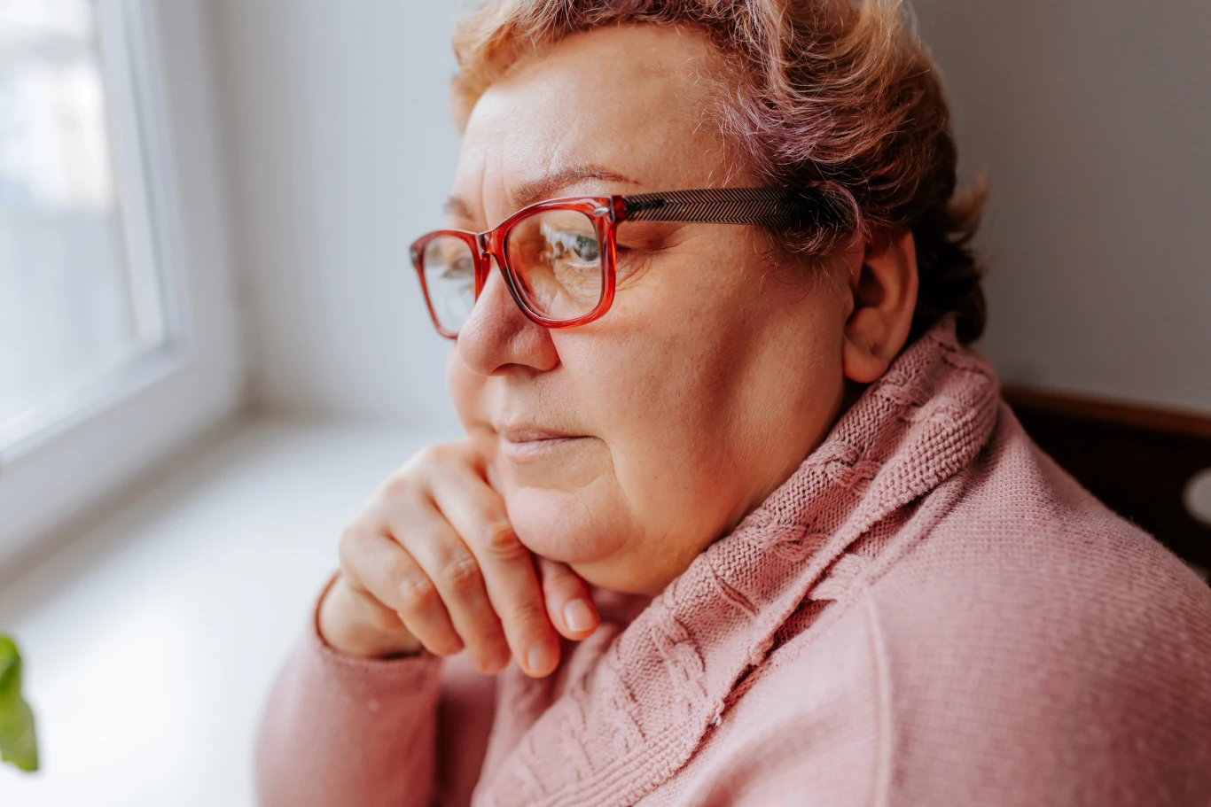 Coraz częściej brakuje ci słów? Nie zawsze jest powód do niepokoju Starsza kobieta w okularach i różowym swetrze opierająca głowę na dłoni, spoglądająca przez okno z zamyśloną miną.