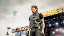 Wokalista w czarnej kamizelce i okularach przeciwsłonecznych, trzymający mikrofon, występuje na dużej scenie festiwalowej podczas koncertu na świeżym powietrzu.