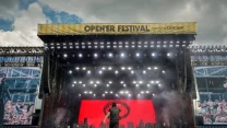 Artysta na scenie podczas koncertu Opener Festival, dynamiczne światła oświetleniowe, duży tłum zgromadzony przed sceną oraz czerwony ekran z graficznym motywem miasta w tle.