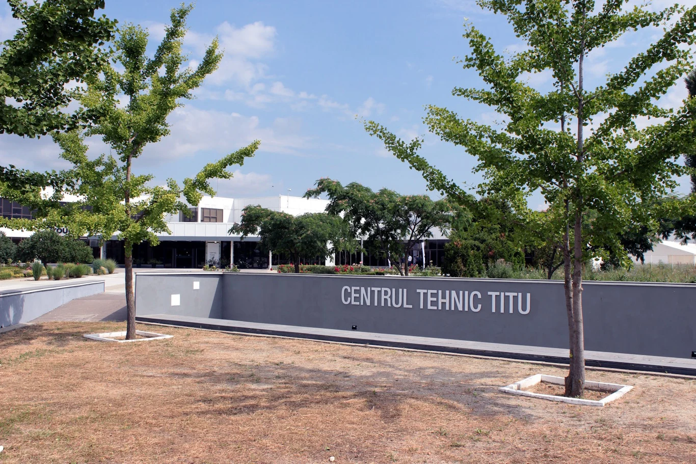 Centrum testowe Grupy Renault w Tiutu Nowoczesny budynek techniczny otoczony zielenią, z wyraźnym napisem 'CENTRUL TEHNIC TITU' na szarej ścianie frontowej i młodymi drzewami wzdłuż alei.