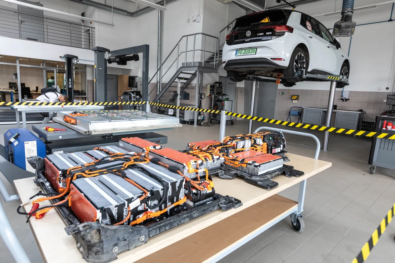 SoH baterii elektryka jest ważniejszy niż przebieg samochodu Akumulatory samochodu elektrycznego na stole warsztatowym, w tle biały auto na podnośniku w serwisie.