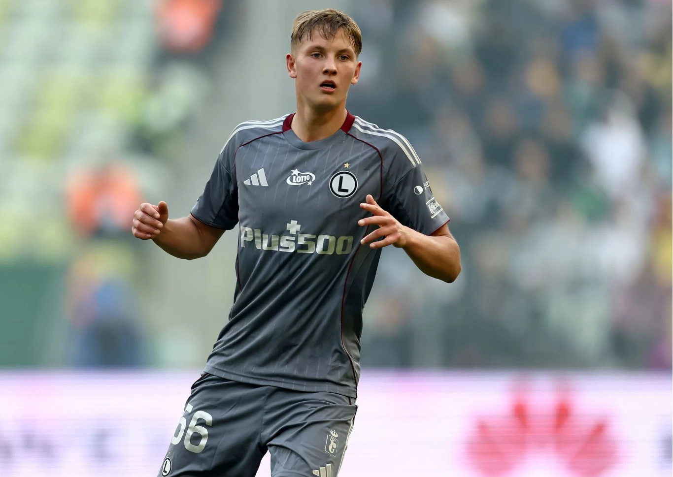 Piłkarz w szarym stroju Legii Warszawa podczas meczu, w dynamicznej pozie na murawie stadionu.