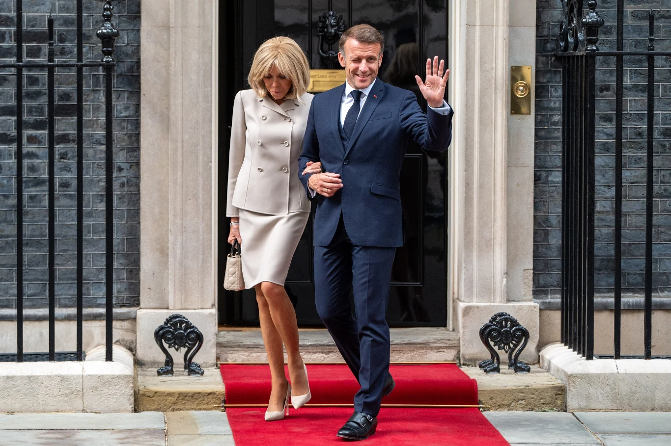 Elegancko ubrana para wychodzi razem z budynku po czerwonym dywanie; mężczyzna w garniturze uśmiecha się i macha ręką, kobieta ma jasny kostium i trzyma torebkę.