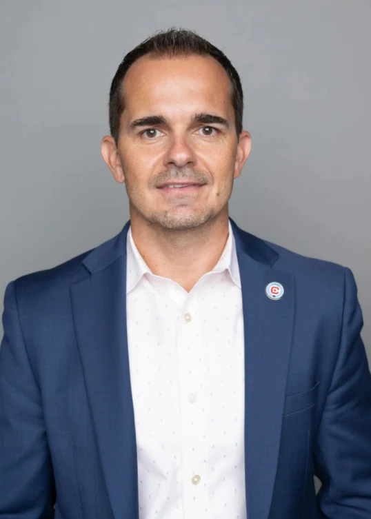 Mężczyzna w niebieskiej marynarce, białej koszuli i z przypinką na klapie, patrzący prosto w obiektyw na neutralnym, szarym tle.
