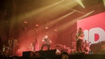 Zespół muzyczny występujący na dużej scenie przy efektownym oświetleniu, z widownią oddzieloną barierkami i dużym ekranem LED wyświetlającym jasny napis w tle.